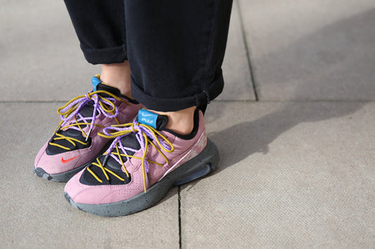 A pair of Nike Air Max Viva in Plum Dust worn with rolled black jeans, laced with purple and mustard cord laces, with a blue Nike Air tab on the heel.