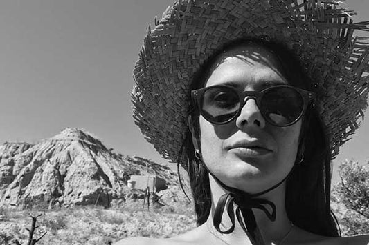 Black and white photo of Zaza de la Hey in a wide woven straw hat and round sunglasses, with a rocky desert hill behind her.