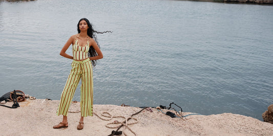 Woman in Damson Madder striped tank top and cropped trousers, brown sandals, standing on a stone quay by calm water.