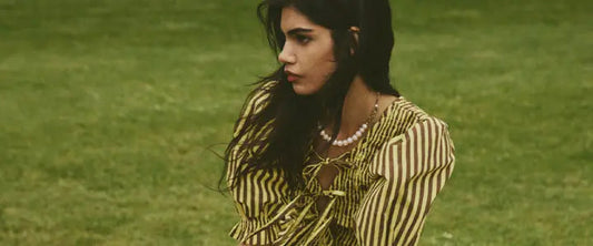 Model in an olive and cream striped Damson Madder blouse with tie fastenings and a pearl necklace, sitting on grass