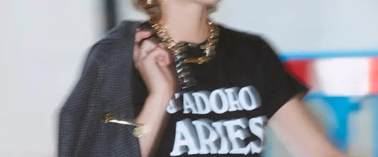 Model in a black Aries J'Adoro Aries slogan t-shirt with a gold chain necklace and a grey checked jacket over one shoulder