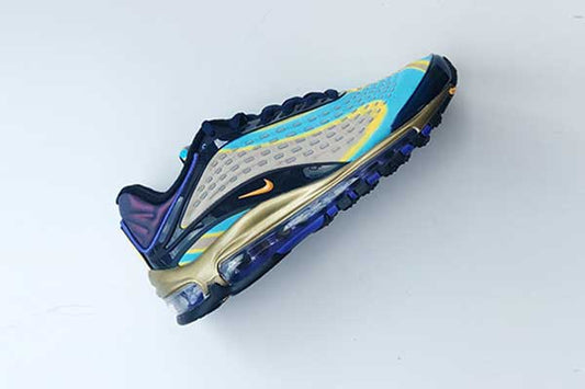 Women's Nike Air Max Deluxe in Midnight Navy/Laser Orange from above, with a light blue and yellow wave-pattern neoprene upper, orange Swoosh, gold midsole, purple iridescent heel and full-length Air unit, on a pale blue background.