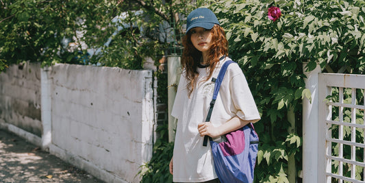 Woman in a Kappy oversized white logo T-shirt, navy cap and blue-and-purple shoulder bag, standing by a leafy fence on a sunlit path.