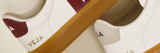 Close-up of two white leather Veja trainers with a burgundy V logo, gold heel lettering and tan rubber soles