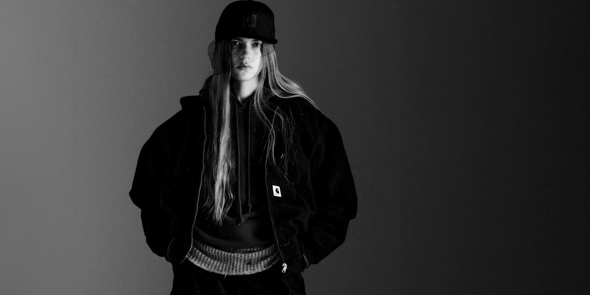 Woman wearing a Carhartt WIP black jacket and cap against a gray background