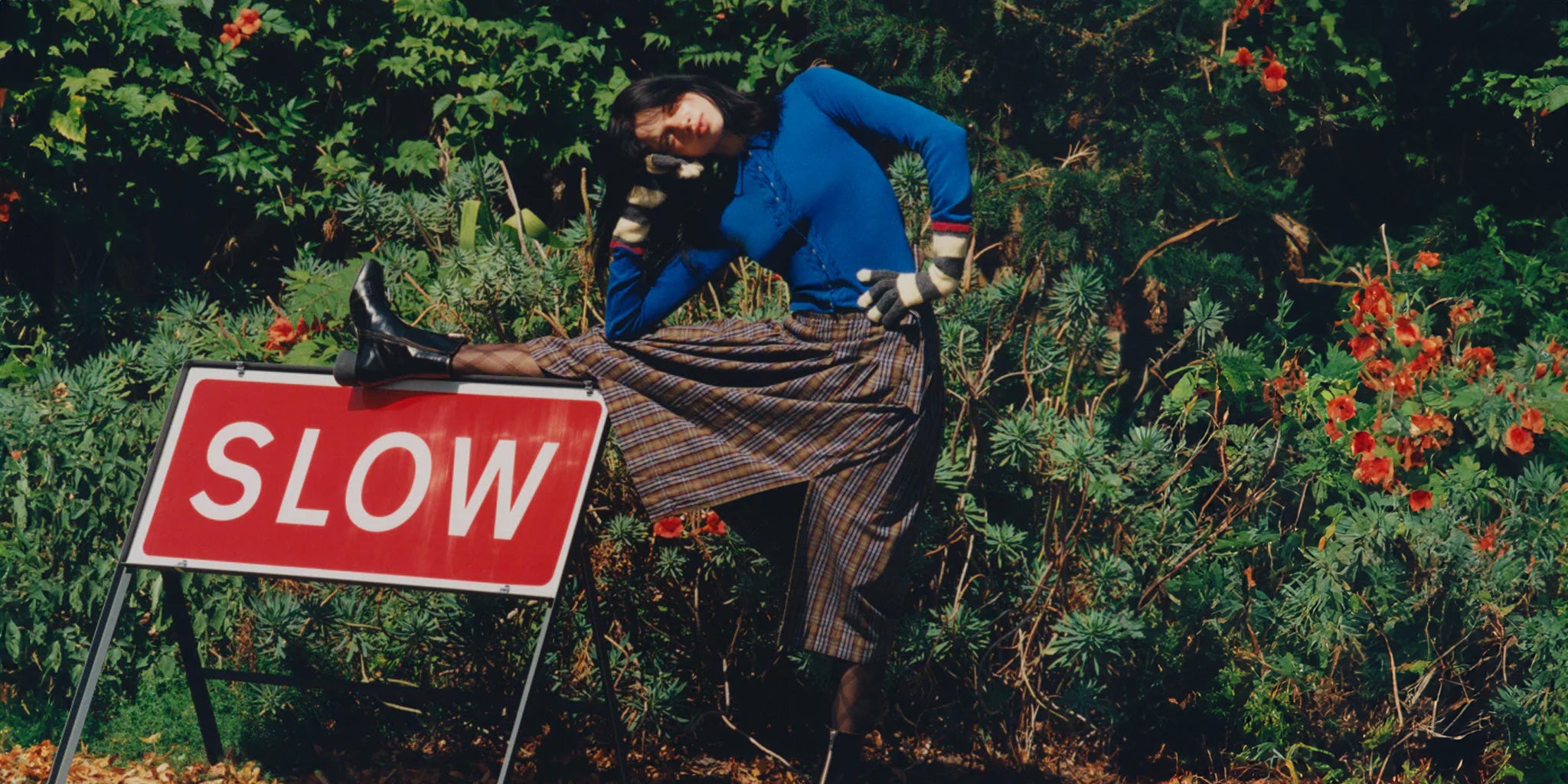 Female wearing a Damson Madder long skirt and blue top standing next to a 'SLOW' sign in a forest setting.